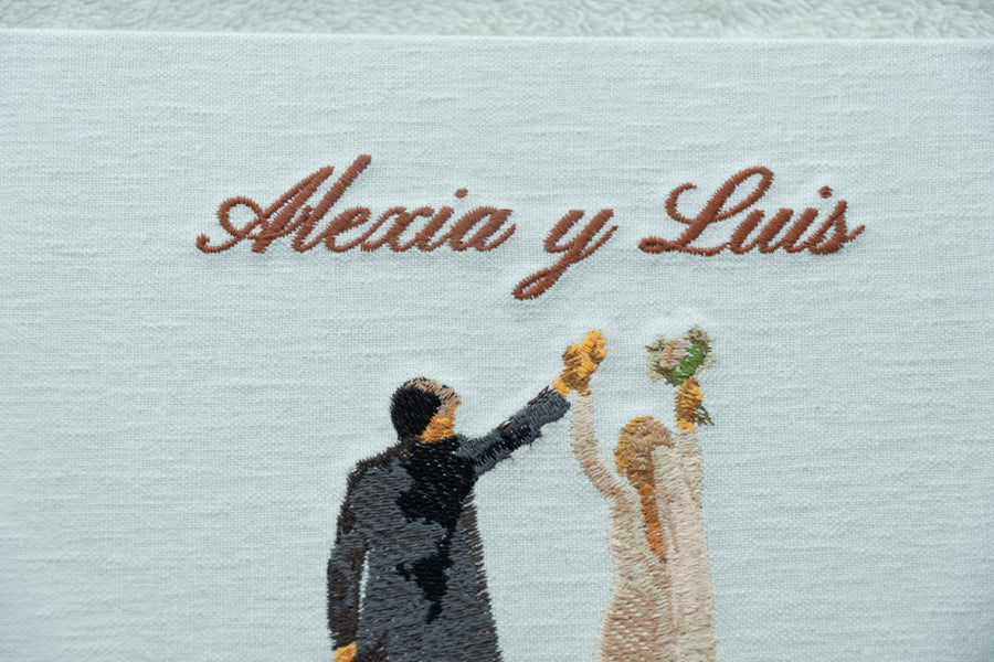 Detalle de la tapa de lino de un álbum de boda bordado a mano con los nombres de la pareja y un dibujo de los novios celebrando